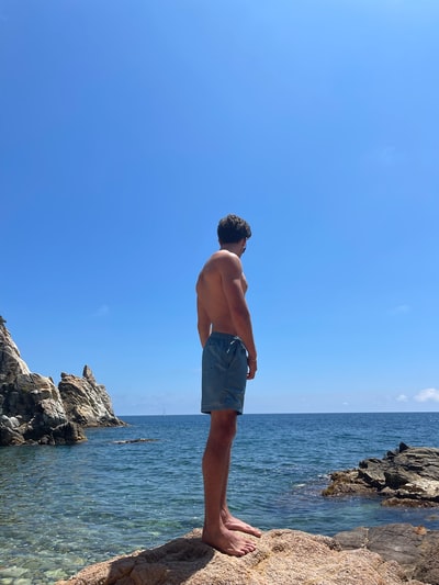 topless man in blue shorts standing on rock formation near body of water during daytime